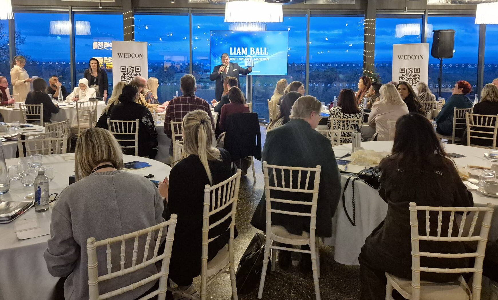 Professional event host presenting to corporate audience using large screen display in modern conference venue