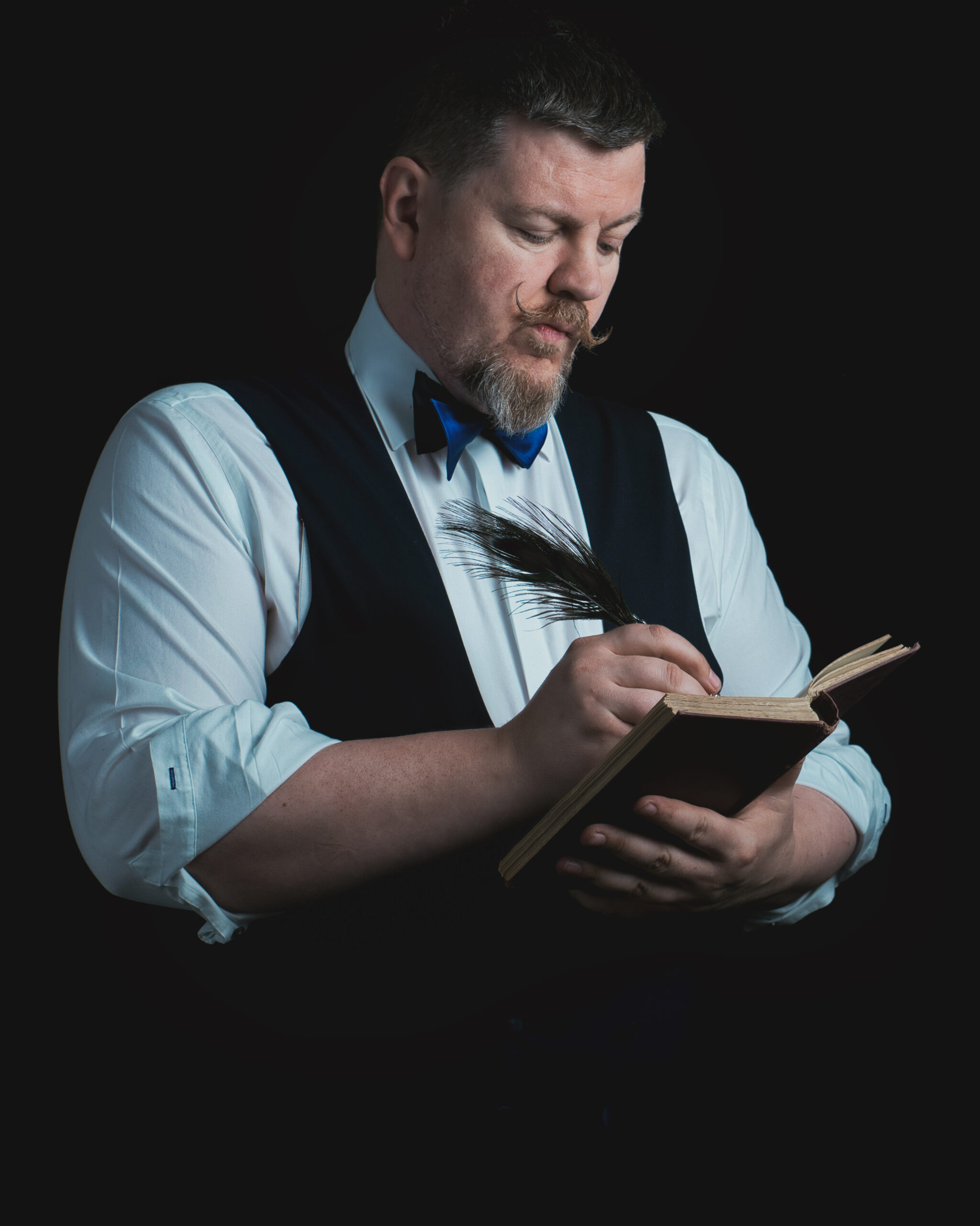 Liam Ball, The Gentleman Magician, dressed in a luxury waistcoat and bow tie, writing with a quill feather in an antique journal against a dark background.