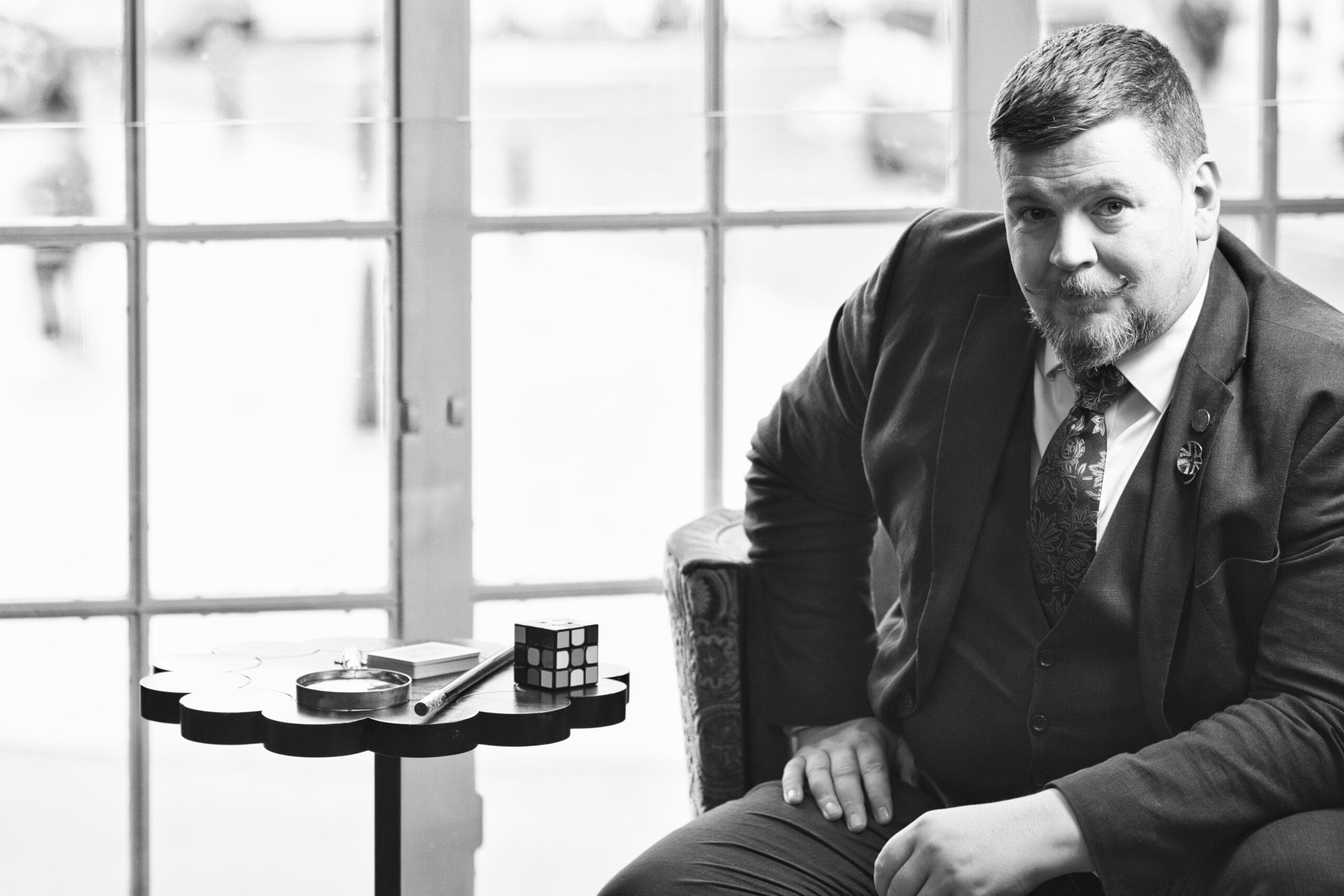 Black and white portrait of Liam Ball, The Gentleman Magician, sitting at a table with a Rubik's cube and playing cards at a venue.
