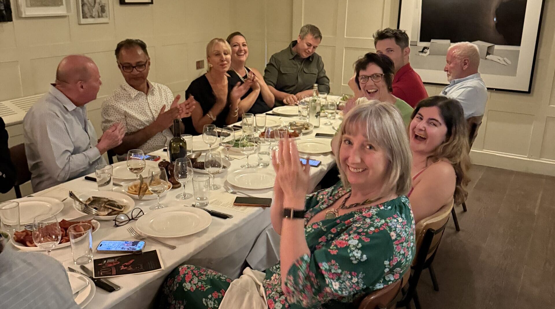 Guests laughing and applauding along a long white dining table during a private event in West London